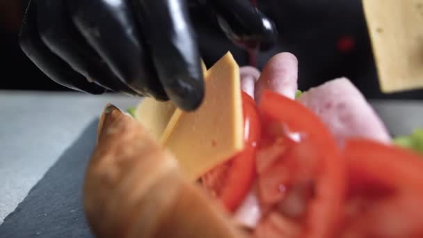 Primer plano de las manos de un chef con guantes negros preparando un sándwich de croissant. Desayuno rápido. Coloque el queso cheddar en el tomate en el tocino a granel. — Vídeos de Stock