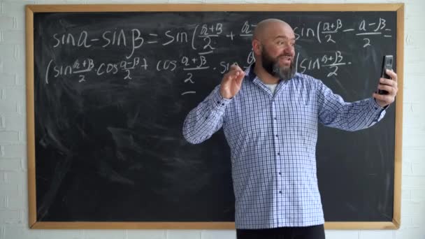 Vuxen manlig lärare tittar på kameran i sin smartphone under en matematik eller algebra lektion. En skäggig och skallig lärare ger en levande lektion. vy från webbkameran. — Stockvideo