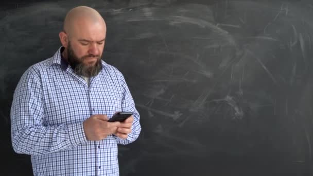 Sakallı beyaz bir adam okulun tahtasının yanında duruyor. Akıllı telefonlu öğretmen. Sosyal ağlarda iletişim, internet bağımlılığı. Boşluğu kopyala. — Stok video