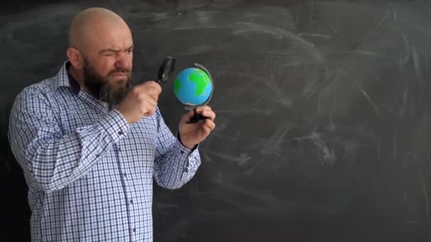 Adulte barbu chauve homme caucasien se tient près du tableau de craie de l'école. Tenez un globe entre vos mains. Verre grossissant. chercher une place sur la carte. espace de copie. — Video