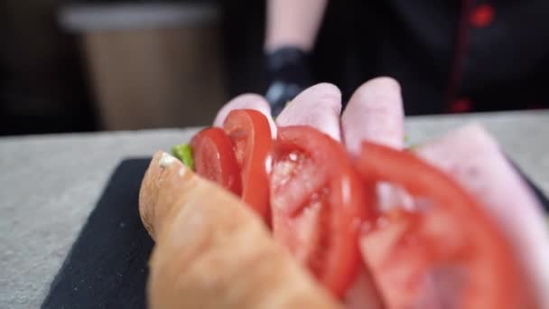 Κοντινό πλάνο των σεφ χέρια σε μαύρα γάντια προετοιμασία ενός σάντουιτς κρουασάν. Γρήγορο πρωινό. Τοποθετήστε τη σφήνα ντομάτας στο ρολό μπέικον. — Αρχείο Βίντεο