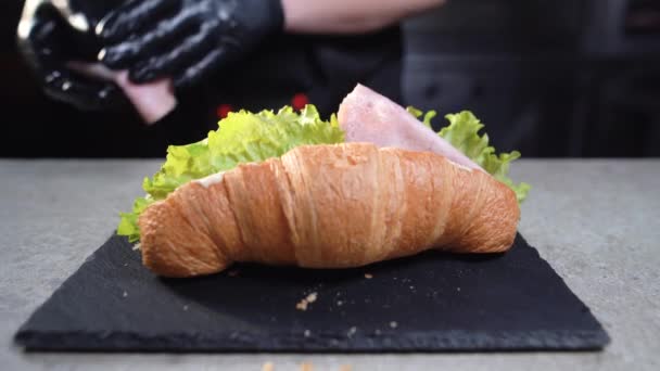 Primer plano de las manos de un chef con guantes negros preparando un sándwich de croissant. Desayuno rápido. Toma tocino y ponlo en un bollo.. — Vídeo de stock