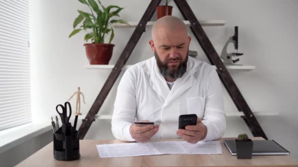 Ernstige dertiger jaren mannelijke arts in een witte jas zit en betaalt voor online bestelling per kaart en mobiele telefoon via het mobiele netwerk in het thuiskantoor. Fysiotherapeut vrijdag. Concept van aankoop op afstand. — Stockvideo