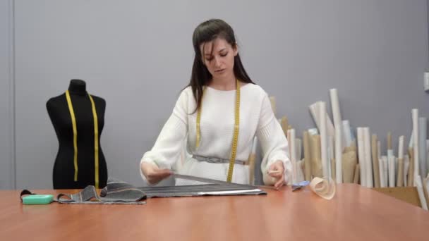 Jonge Kaukasische meisje naaister staat in de buurt van de tafel en is bezig met stof snijden. Concept van designerkleding tijdens het werk. — Stockvideo