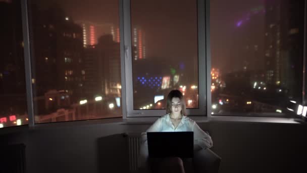 Akşamları evde dizüstü bilgisayarla tek başına oturan beyaz bir kadın. Gece geç saatlere kadar çalışan genç bayan şehir ışıklarına karşı çalışıyor.. — Stok video