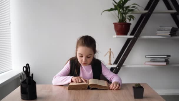 Menina da escola sentada dentro de casa e lendo um livro velho grosso rapidamente virar as páginas. Uma criança gentil e pensativa aprende online remotamente em casa, completando as tarefas dos professores.. — Vídeo de Stock