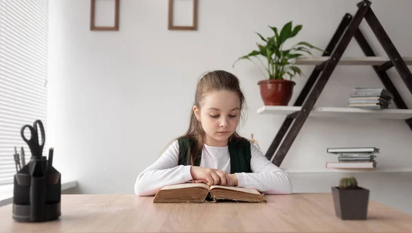 Écolière assise à l'intérieur et lisant un vieux livre épais tournant rapidement les pages. Un enfant délicat et coûteux apprend en ligne à distance à la maison, complétant les devoirs des enseignants. — Photo