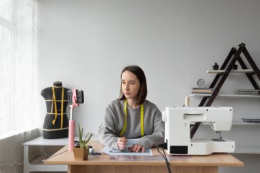 Genç, güzel, beyaz kadın terzi tasarımcılar için eğitim veriyor. Masanın yanına oturur. Çevrimiçi ders, uzaktan eğitim.