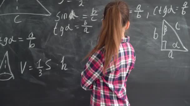 Кавказская школьница в футболке стоит на фоне меловой доски с формулами. пытаясь решить уравнение, улыбаясь. копировальное пространство. — стоковое видео
