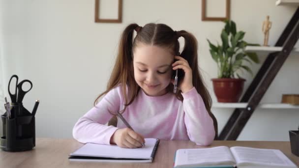 Učitel diktuje domácí úkoly po telefonu, student píše poznámky do sešitu, zatímco sedí u stolu, studuje vzdálenosti on-line. Vzdělávací koncept s pomůckami a mobilní sítí. — Stock video
