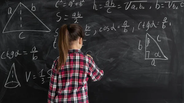 シャツを着た女子学生が黒板に立って数式を書いています。学校に戻るという考えです。スペースのコピー. — ストック写真