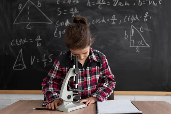 学校に戻ると幸せな時間。黒板の前の教室で勉強している女子高生や学生です。白人の少女は顕微鏡を通して見る。コピースペース. — ストック写真