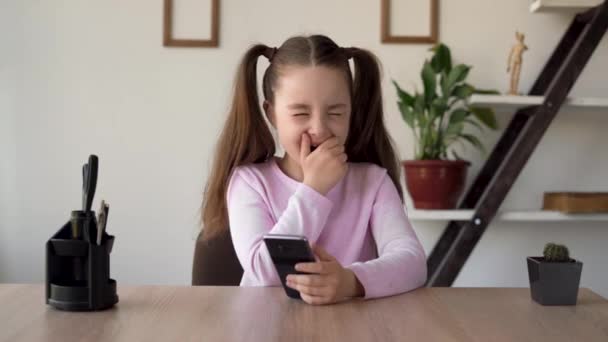 La jeune femme ennuyée et non motivée assise avec un téléphone dans les mains à la table bâille et s'endort. Le problème d'un enfant assis sur les réseaux sociaux et jouant à des jeux en ligne sur un smartphone. — Video