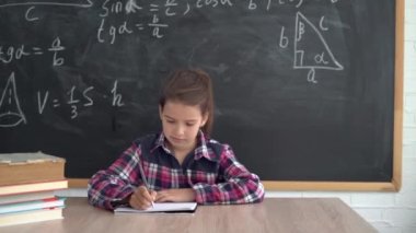 Gömlekli beyaz kız öğrenci formüllü tebeşir tahtasının arka planında oturuyor. Defterine kalemle yazıyor. Okula geri dön..
