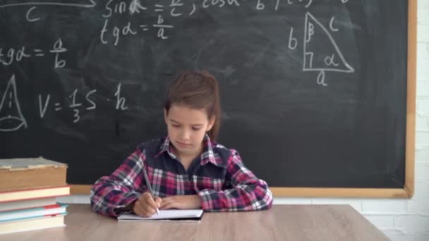 Blank meisje schoolmeisje in een shirt zit aan een tafel tegen de achtergrond van een krijtbord met formules. Hij schrijft met een pen in een notitieboekje. Terug naar school. — Stockvideo
