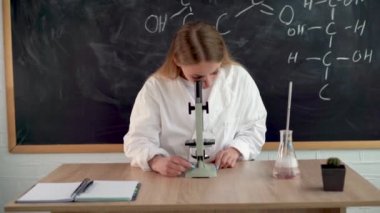 Ciddi bir kız kimyager kolej sınıfında mikroskoptan bakan ve kayıt tutmak için not defterine veri yazan öğrenciler için araştırma yapıyor. Kimyasal deneyler ve araştırma konsepti.