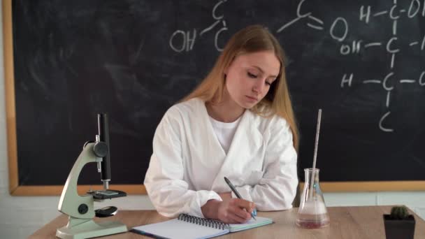 Egy magabiztos kémiatanár egy 25 éves, fehér köpenyes fiatal lány ül egy irodában, és jegyzeteket ír egy krétával, kémiai képletekkel és számításokkal a háttérben.. — Stock videók