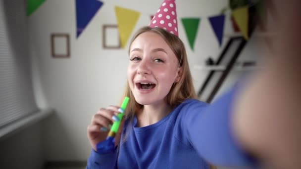 Feliz cumpleaños celebración en línea. Una joven hermosa está celebrando en vivo. gorra festiva en la cabeza. soplando una pipa y sonriendo alegremente. Vista de cámara web. — Vídeo de stock