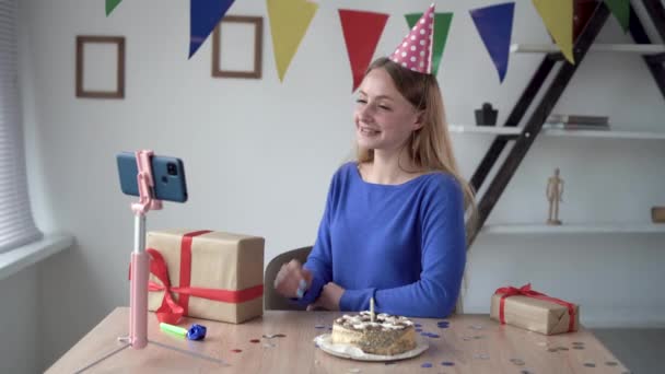 Blondýnka video blogger pozdravuje své předplatitele ve vysílání. narozeniny online. doma u stolu na dálku. Chytrý telefon je na stativu.. — Stock video