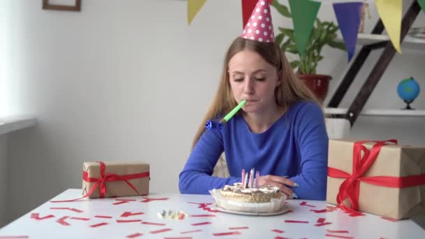 Eine langweilige und traurige Geburtstagsfeier. Eine blonde Dame sitzt zu Hause am Tisch, betrachtet die Torte und bläst in eine Pfeife. Schachtel mit roter Schleife. — Stockvideo