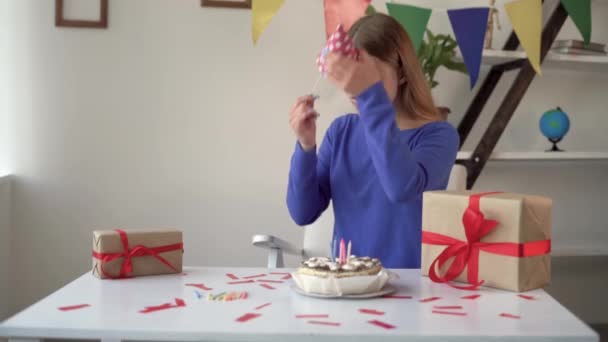 Una rubia caucásica está sentada en casa en la mesa. Se pone una gorra festiva en la cabeza. Celebrando un feliz cumpleaños solo. — Vídeos de Stock
