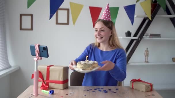 Fiesta de cumpleaños en línea. Una mujer caucásica se sienta en una mesa en casa y transmite su cumpleaños a través de la aplicación. Él toma el pastel en sus manos y sopla las velas. — Vídeos de Stock