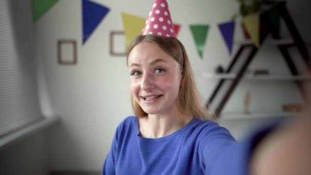 A menina branca loira em um gorro festivo celebra um aniversário. Ele transmite on-line usando um smartphone e através de um aplicativo. Mostra uma caixa com um arco vermelho. — Vídeo de Stock