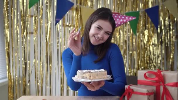 Glückliche 20-jährige Kaukasierin steckt lächelnd eine Kerze in eine Geburtstagstorte und feiert ihren Geburtstag zu Hause mit Dekorationen. Party vor der Webcam und Feier mit Dessert. — Stockvideo