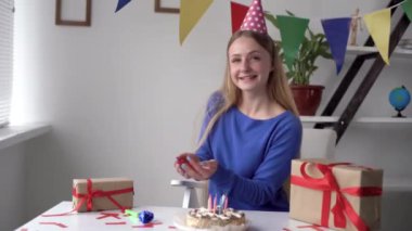 Neşeli bir sarışın Kafkasyalı kız evdeki masada oturuyor. Koltukta dans etmek gibi. Ellerini kaldırdı ve konfetiyi savuşturdu. Doğum günü havai fişekleri. hediyeli kutular.