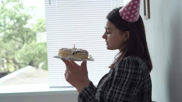 Uma jovem empresária europeia está em um gorro festivo junto à janela com um bolo nas mãos e apaga a vela de aniversário no escritório. Uma mulher com sobremesa no quarto. — Vídeo de Stock