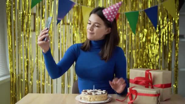 Cumpleaños en vivo en la cámara del teléfono de una chica alegre cuenta y muestra regalos y un pastel de cumpleaños. Una señora con un sombrero festivo se sienta a la mesa en casa. Parte concepto de distancia. — Vídeos de Stock