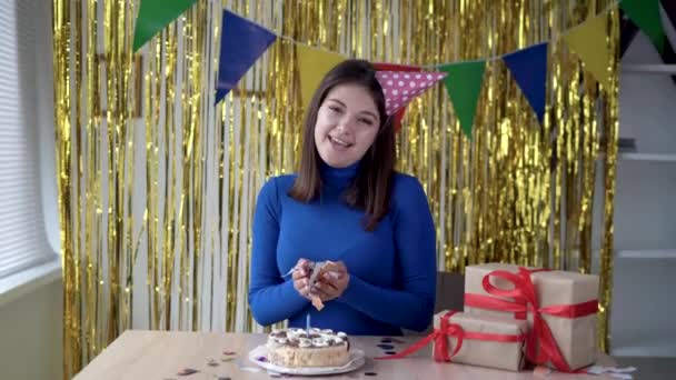 Cumpleaños de las mujeres en la cámara web, una chica con un sombrero festivo sonríe y habla de la fiesta, vomita confeti de colores. Señora sentada a la mesa con un pastel de cumpleaños y regalos. — Vídeos de Stock