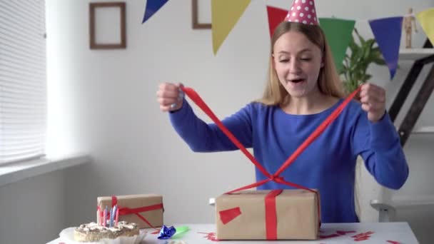 Cumpleaños en casa solo. Una joven sentada en una mesa desata un lazo en una gran caja de artesanía. Señora feliz y alegre cumpleaños. — Vídeos de Stock