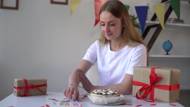 Voorbereiden op een verjaardagsfeestje. Jonge mooie blonde vrouw zit aan de tafel en steekt kaarsen in de verjaardagstaart. — Stockvideo