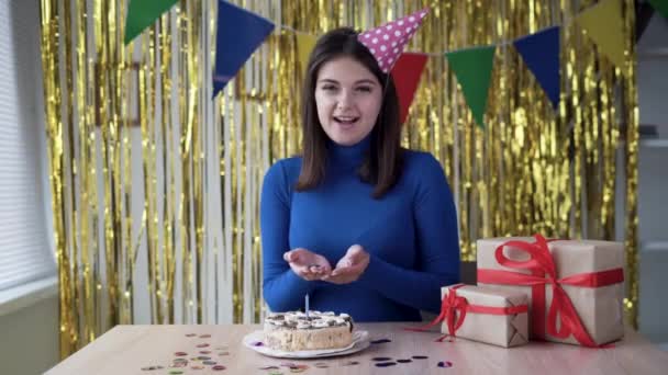 Cumpleaños de una joven chica caucásica en línea en la cámara en una habitación con decoraciones. Mezcle confeti multicolor de la palma de la mano. Concepto remoto de vacaciones. Celebración del aniversario. — Vídeos de Stock