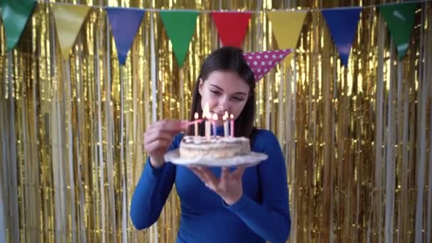 Caucásico millennial enciende velas en pastel de cumpleaños en casa celebrando su cumpleaños. Una chica con un postre en sus manos contra el telón de fondo del paisaje en la habitación. Gorra de fiesta en la cabeza — Vídeos de Stock