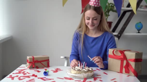 Uma jovem mulher em um gorro festivo senta-se na mesa sozinha em casa. Acende as velas no bolo. O conceito de um aniversário em casa enquanto isolado. — Vídeo de Stock