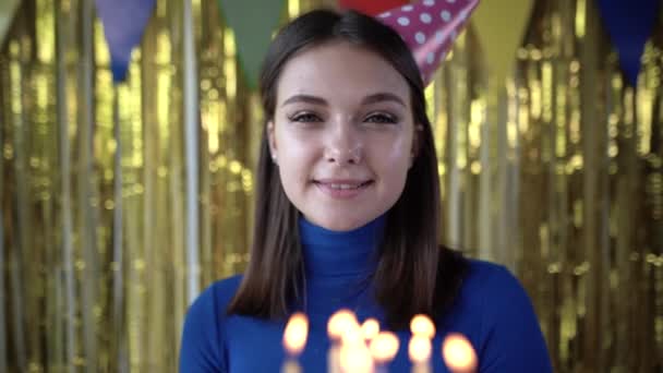 Primer plano de un pastel de cumpleaños con velas encendidas en las manos de una niña en una gorra de cumpleaños de pie en una habitación en casa con decoraciones de oro en el fondo. Concepto de partido en primera persona. — Vídeos de Stock