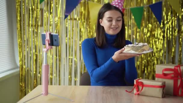 Alegre estudiante bloguera caucásica sentada en la mesa en casa celebra su cumpleaños, mostrando pastel en la cámara del teléfono celular soplando 1 vela. Concepto de fiesta. — Vídeos de Stock