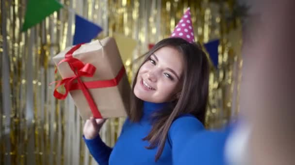 Menina milenar caucasiano feliz mostra uma caixa com um presente em seu aniversário em uma câmera de smartphone. Senhora de chapéu festivo com uma surpresa nas mãos. Vista da câmara. — Vídeo de Stock