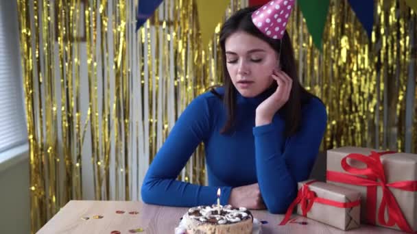 Evde yalnız başına depresyondaki beyaz bir bayanın sıkıcı doğum günü. Kız şenlik şapkası ve pastası içinde bir hediyeyle masada oturmaktan sıkıldı. Tatil dekorasyonları. — Stok video