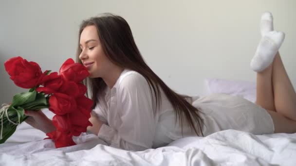 Chica caucásica 20 s en una camisa se encuentra en la cama con un ramo de flores, los huele y disfruta de la mañana. Tulipanes rojos en las manos de una jovencita. Mujer acostada en el dormitorio. — Vídeos de Stock