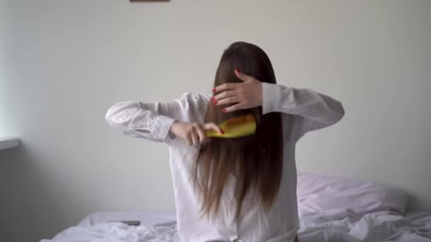 Chica seria de 20 años se peina el cabello con un peluquero mientras está sentado en la cama. Cuidar la salud y la longitud del peine. La prevención del desequilibrio hormonal en la vida de las mujeres. — Vídeos de Stock