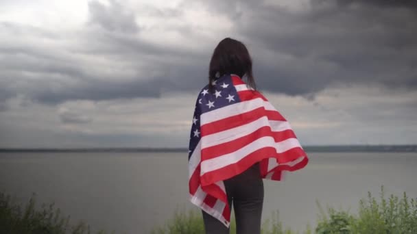 Женщина с американским флагом на плечах стоит на фоне облачного неба с облаками. День независимости Америки и День флага. Копирование пространства. — стоковое видео