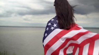 Arkadan yakın plan portre. Omzunda Amerikan bayrağı olan bir kadın bulutlu bir gökyüzünün arka planında duruyor. Amerika bağımsızlık günü ve bayrak günü. Boşluğu kopyala.