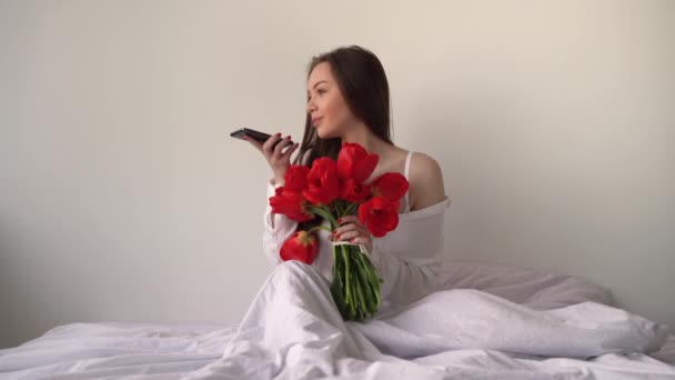 Glückliche Brünette im Hemd mit einem Blumenstrauß auf dem Bett im Schlafzimmer diktiert eine Nachricht im Boten ans Telefon. Handy und Sprachrekorder in den Händen eines jungen kaukasischen Mädchens — Stockvideo