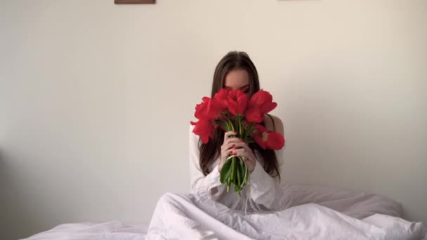 Een dame met een boeket bloemen zit op het bed in de slaapkamer, snuivend aan tulpen en niezende allergieën. Ochtend en millennium op beddengoed en een orthopedische matras. Het concept van de ochtendvrouw. — Stockvideo