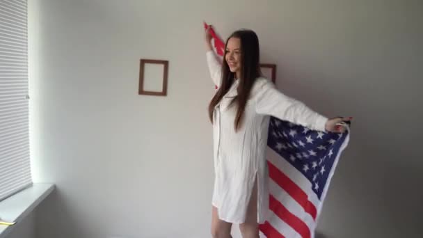 Una hermosa chica caucásica en el Día de la Bandera Americana está bailando en casa en el dormitorio en la cama con una bandera en sus manos. 14 de junio en América. Independencia de Estados Unidos y concepto de bandera. — Vídeos de Stock