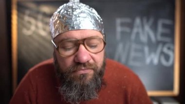 Koruyucu folyo şapkalı, sakallı, beyaz bir adam video blogu yönetiyor. Web kamerasına bakar ve sahte mesajlı bir işaret gösterir. Komplo teorisi kavramı.