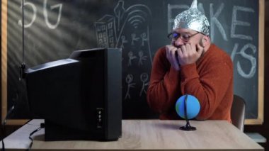 Sakallı bir komplo teoristi evinde masasında oturmuş eski bir TV izliyor. Kafaya folyo şapka takılır. Paranoyak korkuyor. Komplo teorisi kavramı.